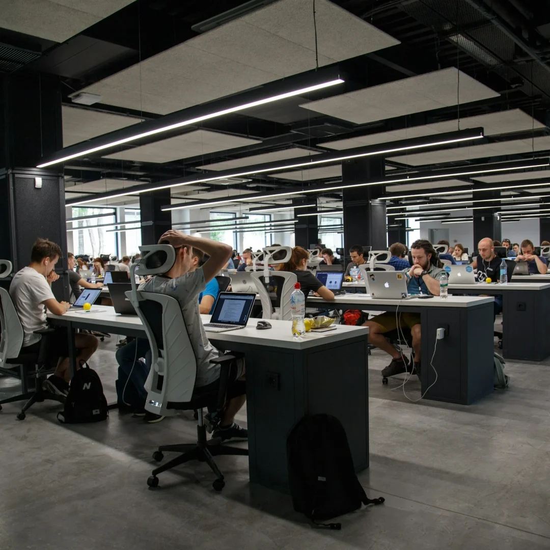 A spacious, modern office with rows of employees working on laptops at long desks. The atmosphere is focused and industrious, with bright lighting.